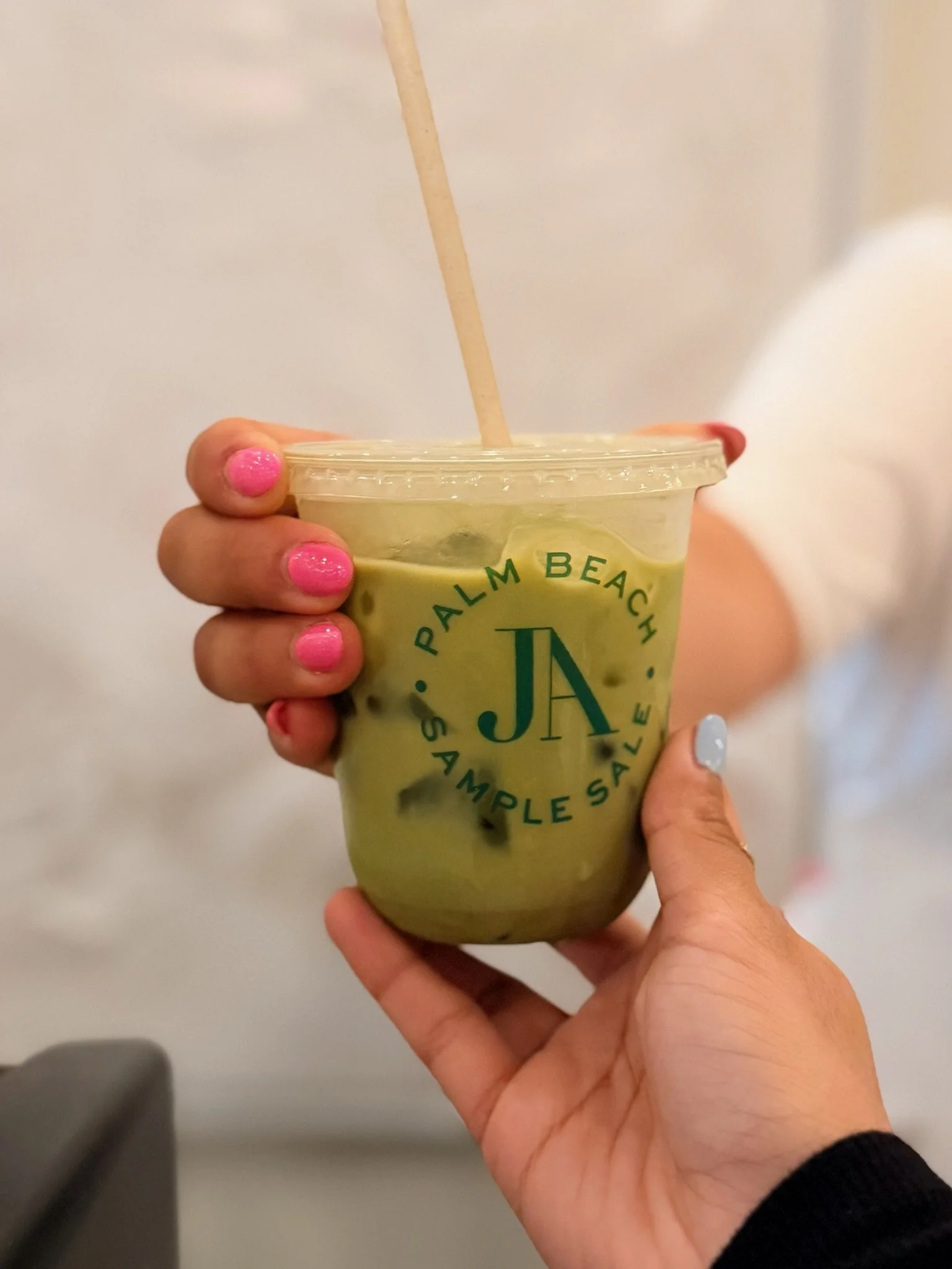 A person holding a plastic cup of green matcha latte with an arm decorated with pink and light blue nail polish. The cup has a logo that reads "Palm Beach" with additional text.