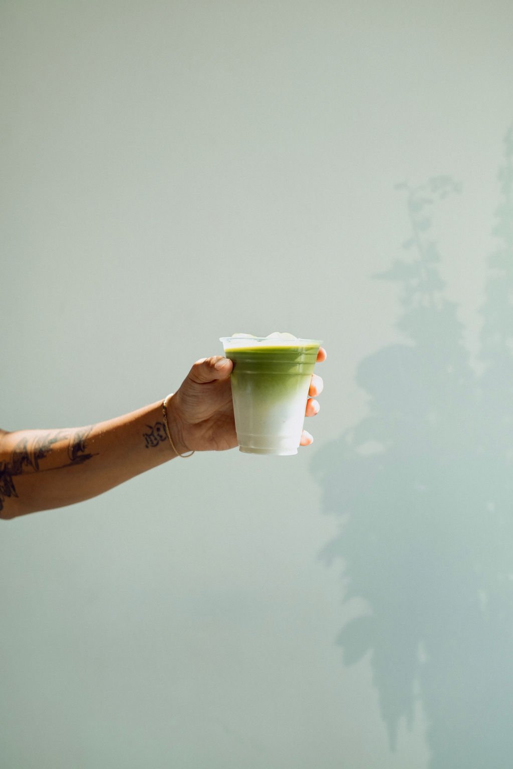 A person's hand holding a glass of green layered drink against a plain light-colored wall.