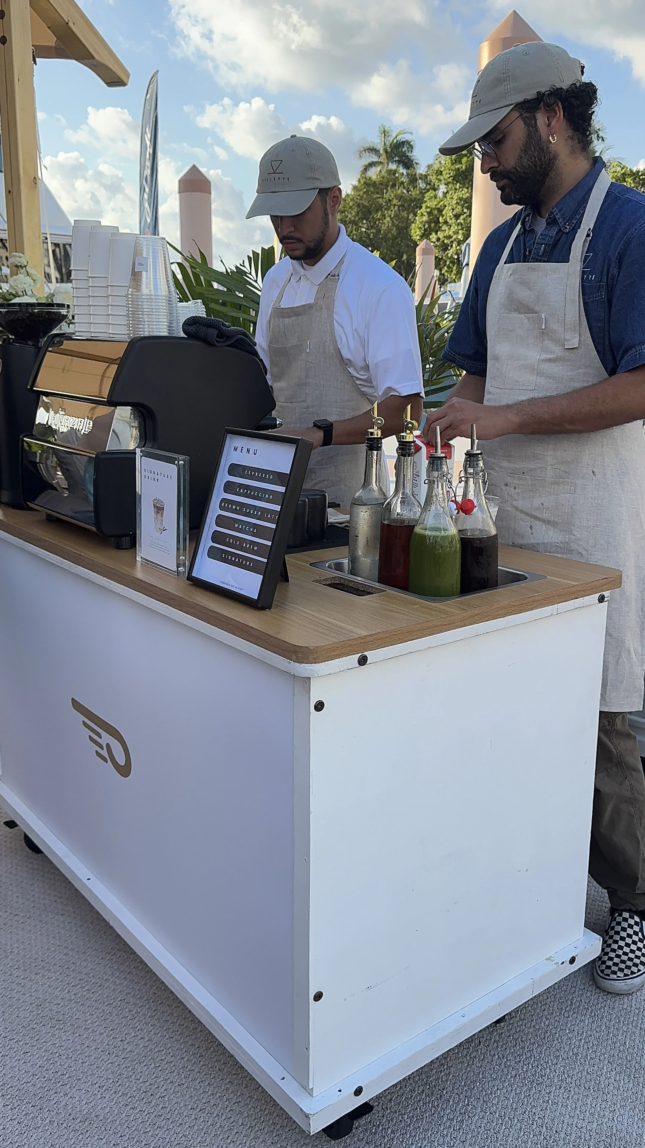 A mobile coffee stand with two baristas preparing drinks, surrounded by drink bottles, a menu, and cups, outdoors under a partly cloudy sky.