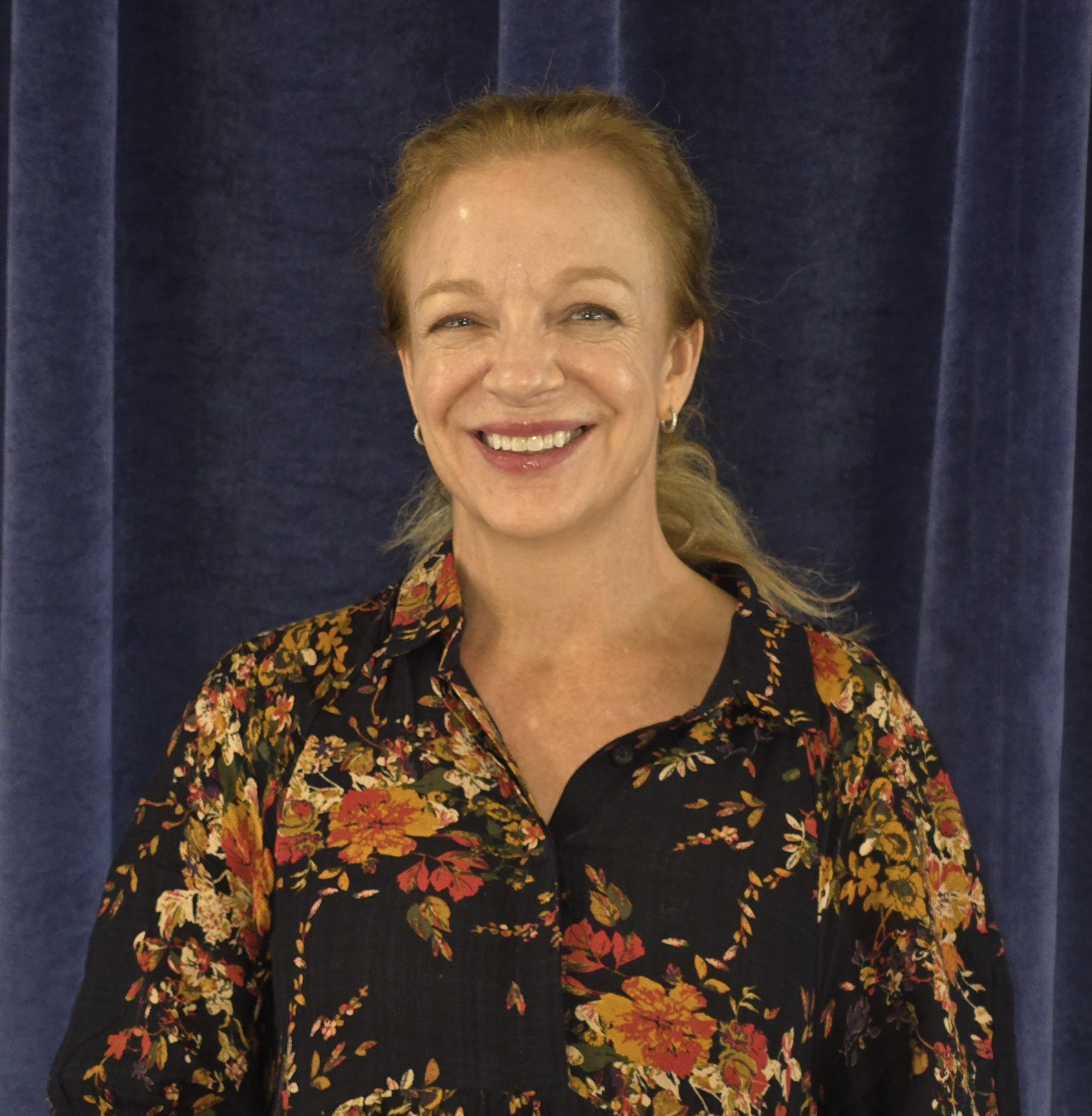 A woman with red hair, wearing a floral patterned blouse and earrings, smiling in front of a dark curtain.