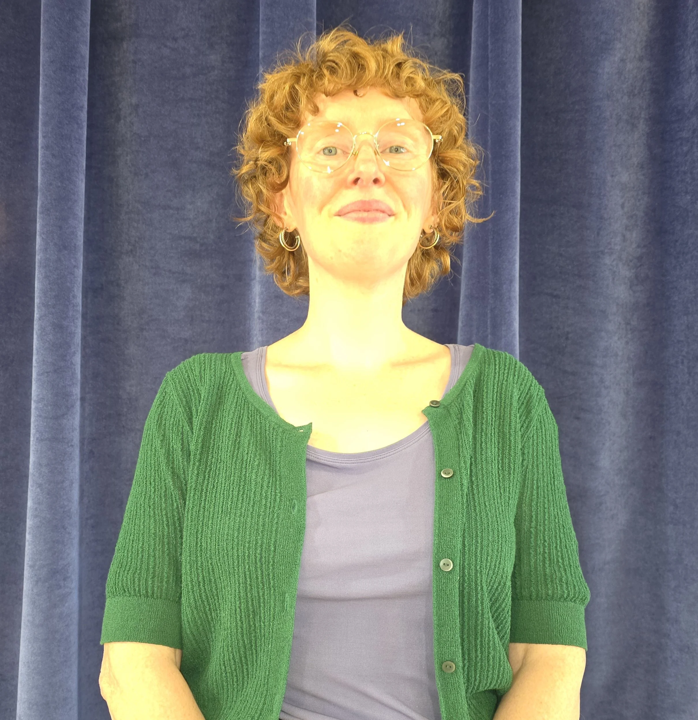 A woman with curly red hair wearing glasses, a gray shirt, and a green cardigan, standing in front of a dark blue curtain.