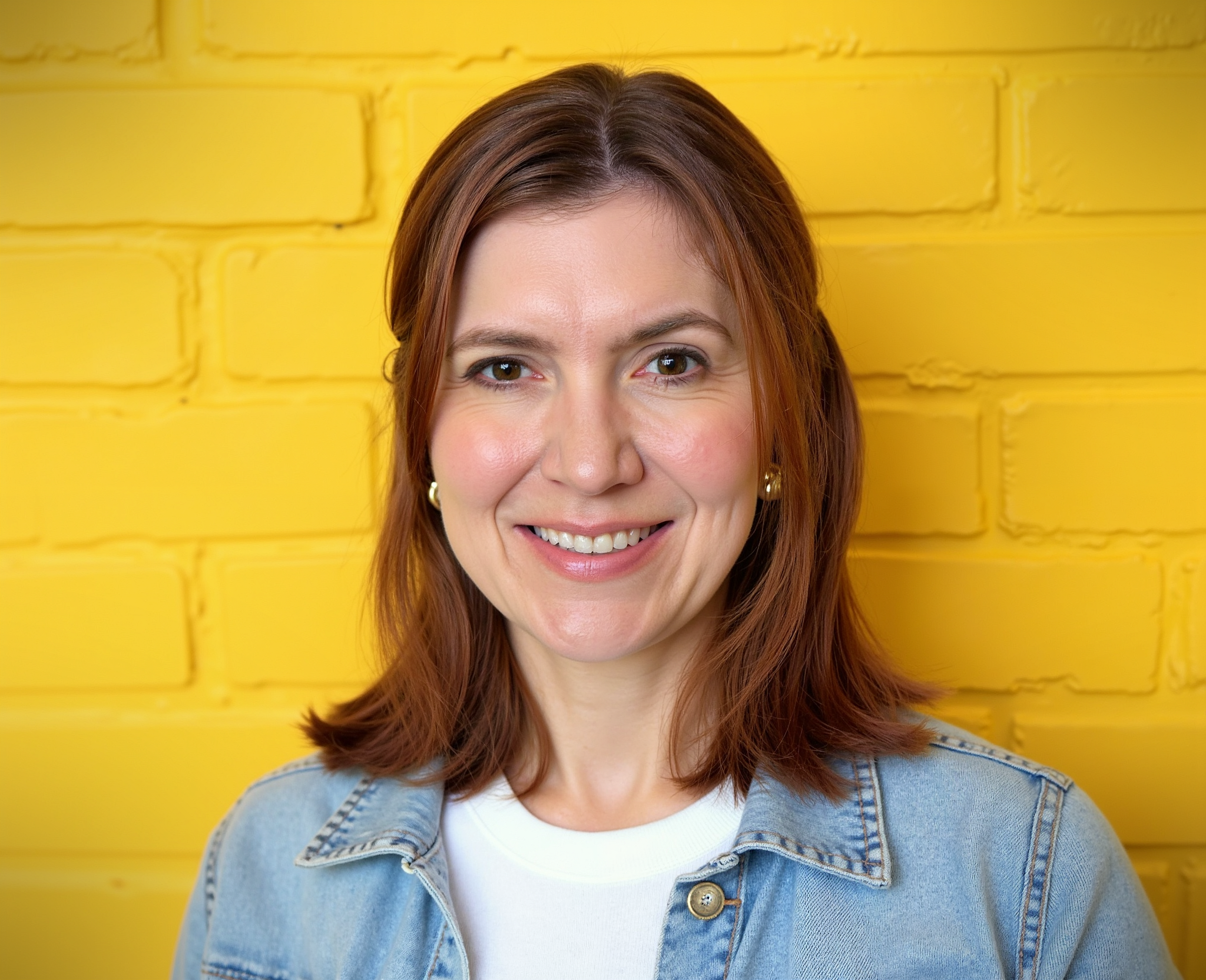 A woman with shoulder-length red hair, smiling, standing in front of a yellow brick wall, wearing a light denim jacket and a white shirt.