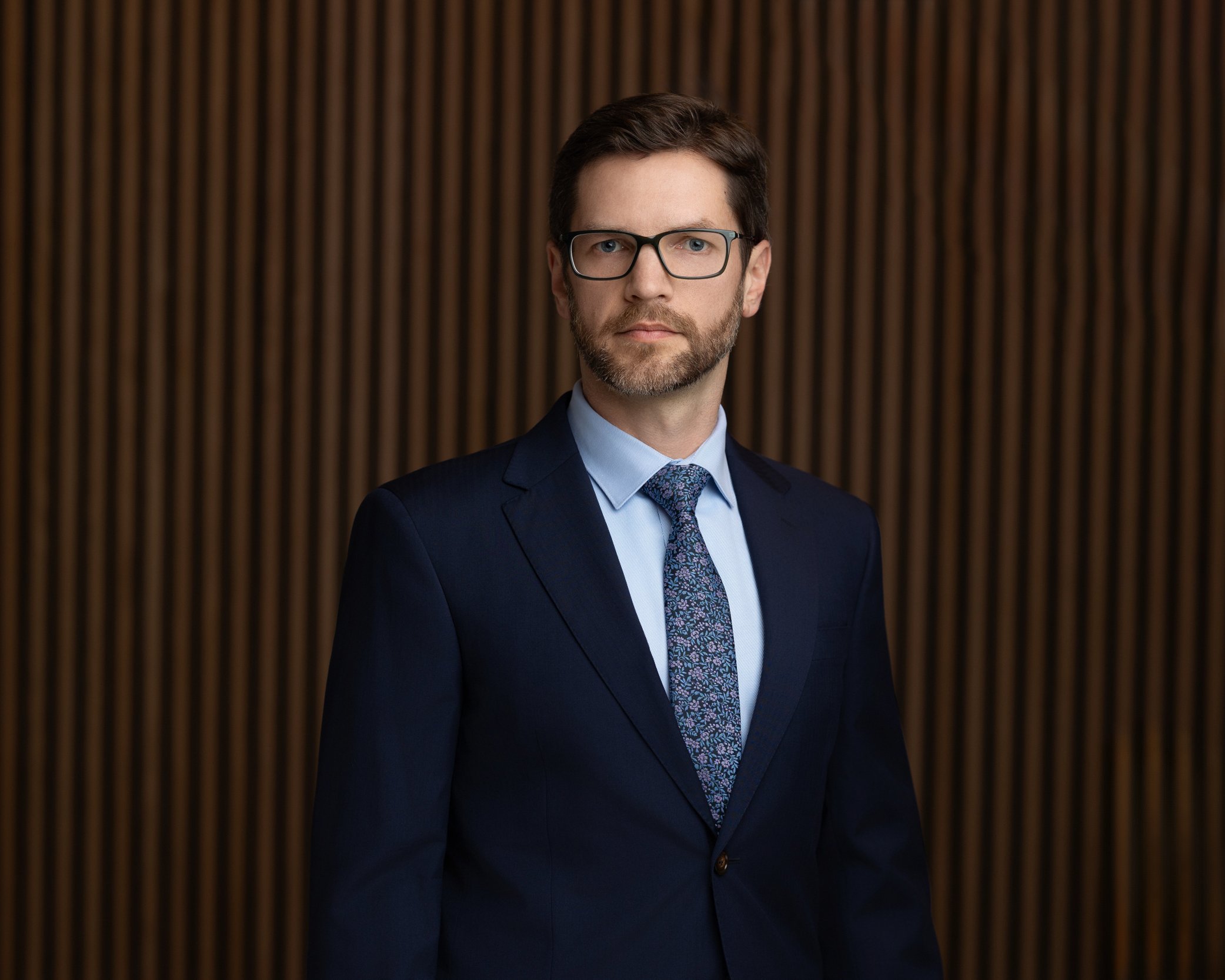 A man with brown hair, beard, glasses, wearing a navy suit, light blue shirt, and patterned tie, standing in front of a wooden paneled wall.