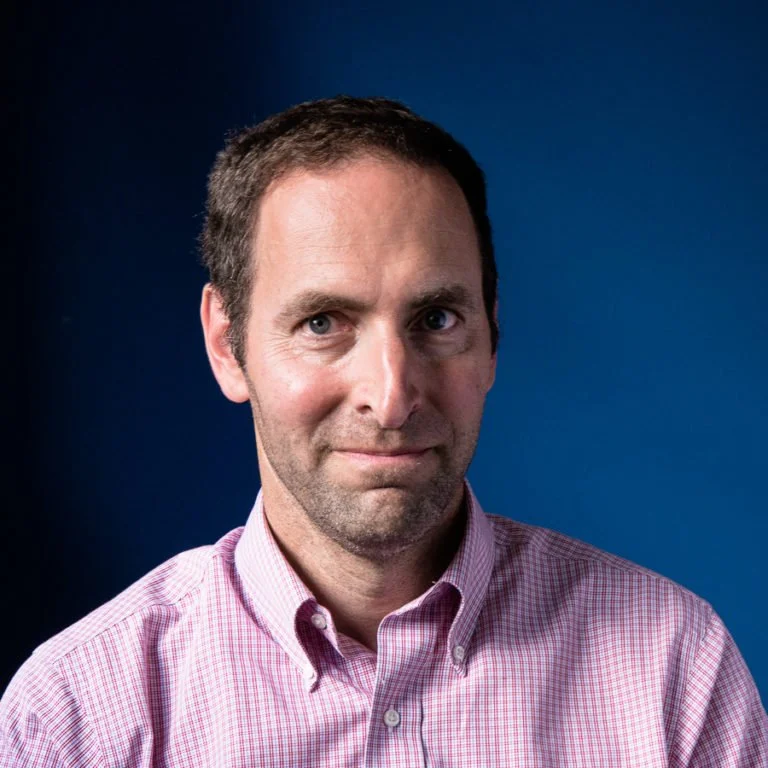 Headshot of a man with short brown hair, wearing a light pink checkered button-up shirt, smiling slightly against a blue background.