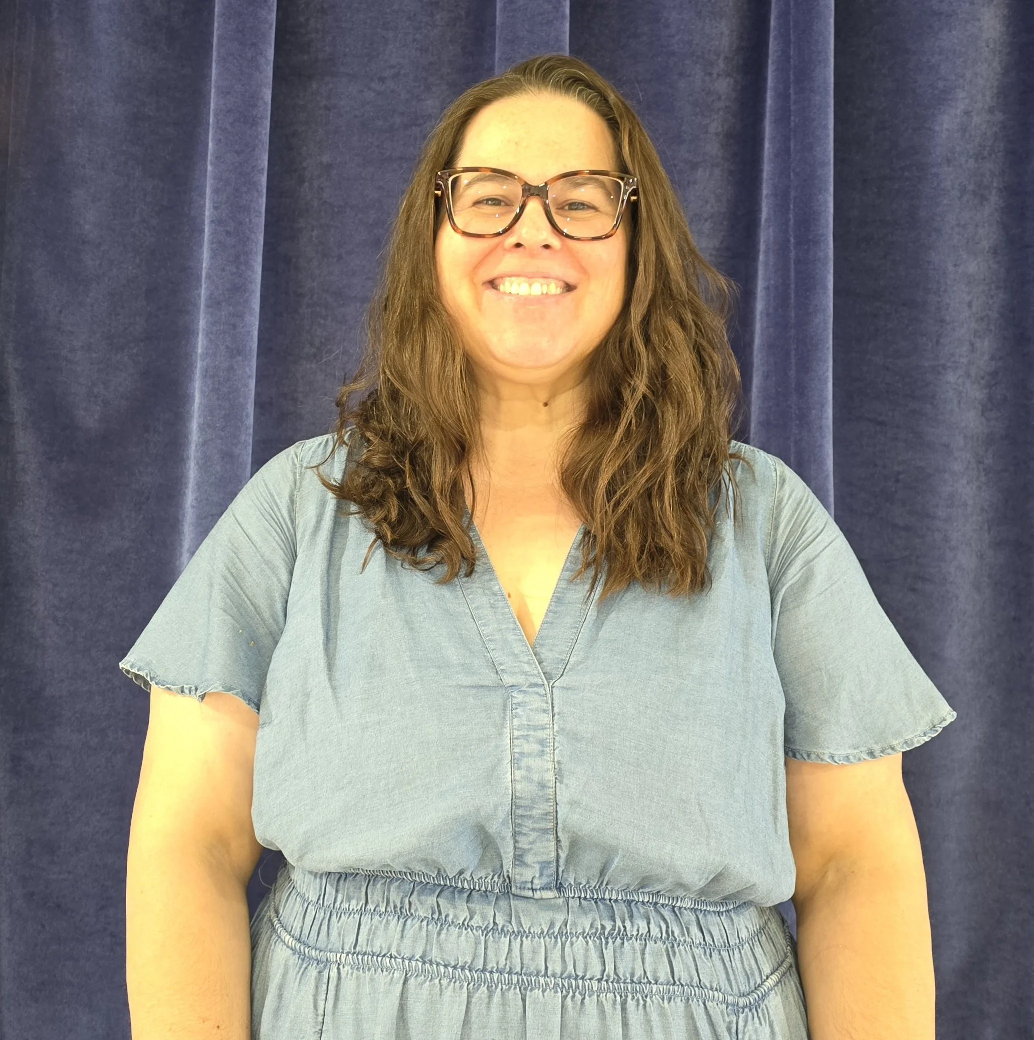 Close-up of a woman with long wavy brown hair wearing large tortoiseshell glasses and a gray shirt standing in front of a blue curtain, smiling.