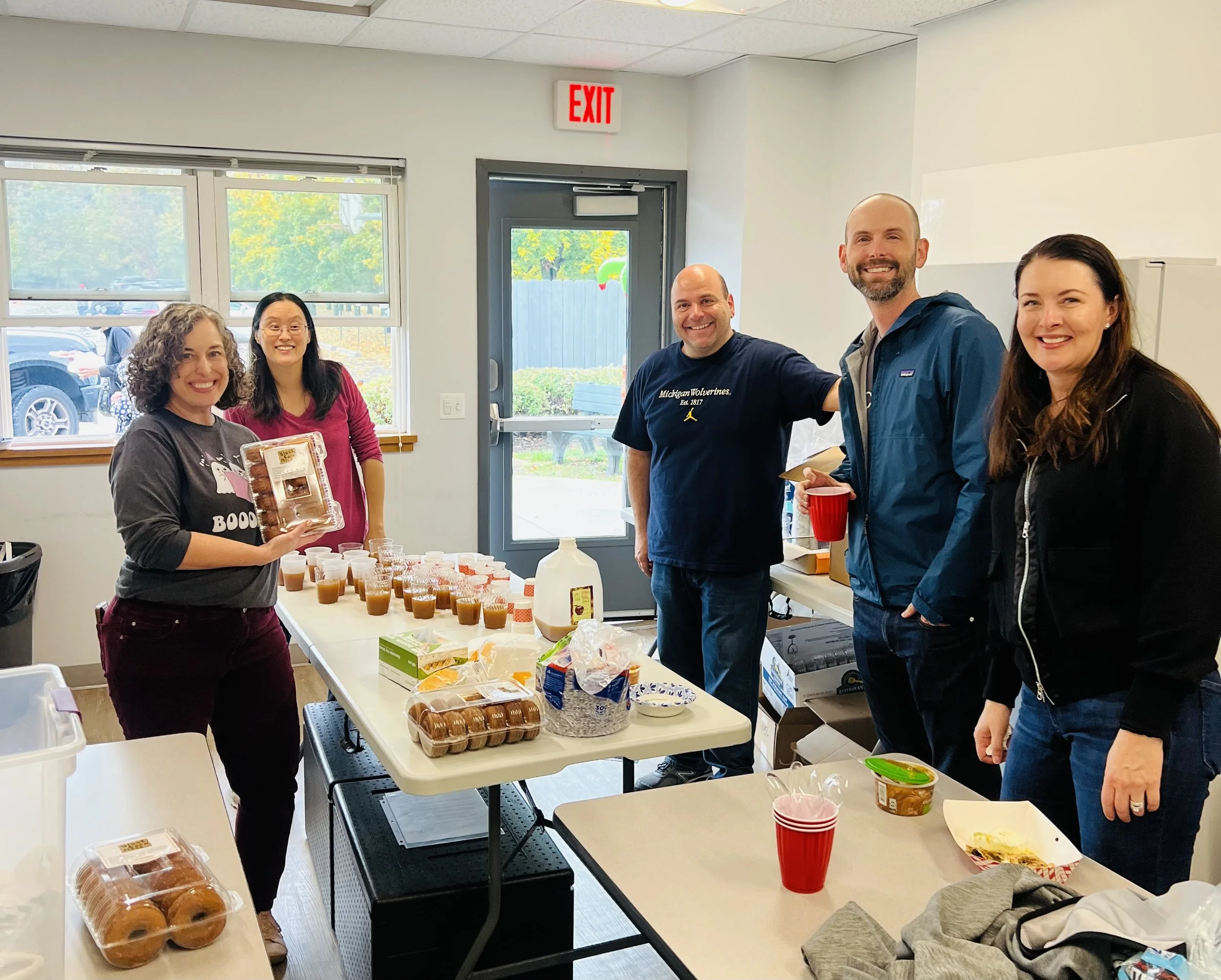 Group of five people standing inside near a table with baked goods, cups, and snacks, smiling at the camera during a gathering or celebration.