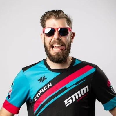 A person with light skin, brown hair, and a beard wearing red sunglasses and a colorful cycling jersey with black, blue, red, and white stripes, and various sponsor logos, smiling and sticking out his tongue against a plain gray background.