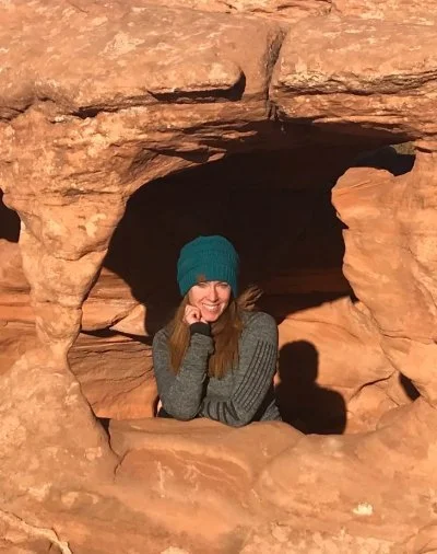 A woman with long red hair, wearing a teal knit hat and a gray long sleeve shirt, smiling and sitting in a natural rock formation with a small opening, in a desert landscape.