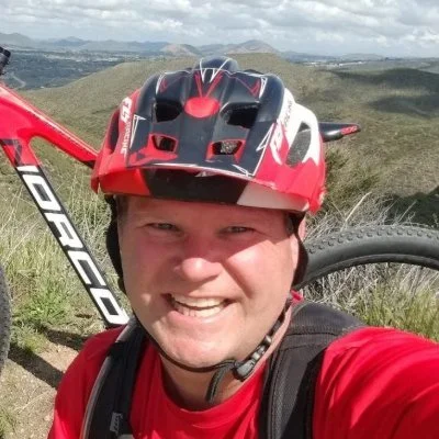 Smiling man with a red and black helmet, taking a selfie with a mountain bike on a trail with scenic hills and cloudy sky in the background.