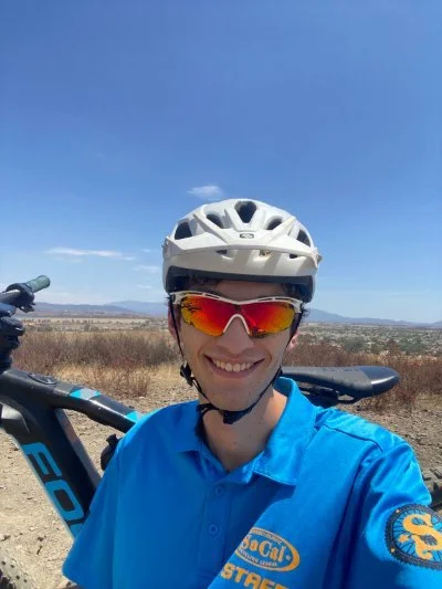 A smiling man wearing a white helmet, orange reflective sunglasses, and a blue cycling jersey with a patch that says 'PCCAI - Cycling League' and 'STAFF'. He is taking a selfie outdoors with a clear blue sky and a flat landscape in the background, along with a mountain range.