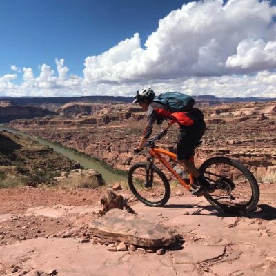 A person riding a mountain bike on a rocky trail at the Grand Canyon, with a river flowing below and a partly cloudy sky overhead.