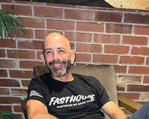 Smiling man with a beard and shaved head, wearing a black T-shirt with white text, sitting in a chair in front of a brick wall.