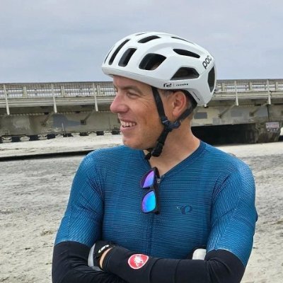 A man at the beach wearing a white bike helmet and a blue athletic shirt with black sleeves. He has sunglasses hanging on his shirt and is smiling while looking to the side, with a pier or bridge in the background.