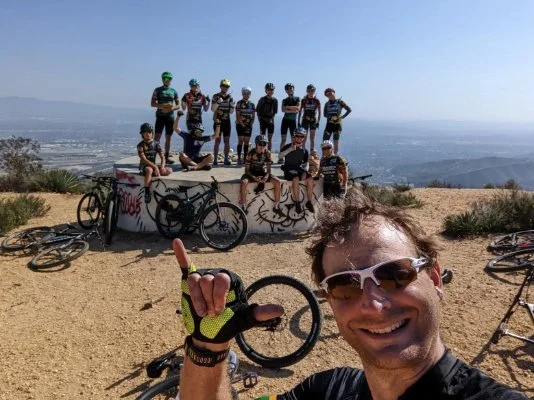 A group of people, including children and adults, wearing cycling gear and helmets, gathered on a large, graffiti-covered circular concrete structure on a hilltop with mountain and valley views in the background. There are several bicycles lying on the ground in front of them, and a man in the foreground taking a selfie, smiling and giving a thumbs-up.