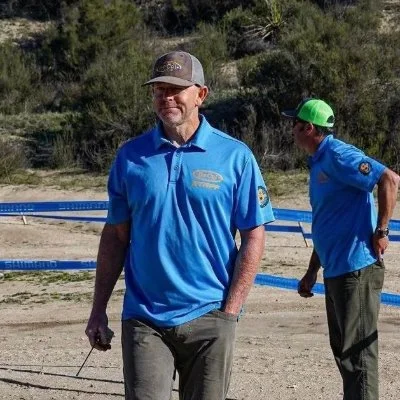 Two men in matching blue shirts and hats outdoors, with a blue fence and green bushes in the background.