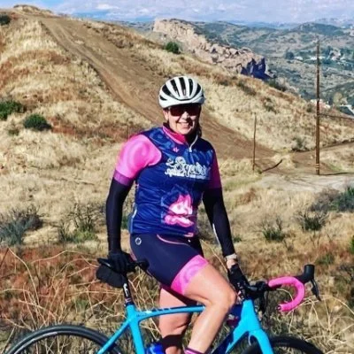 A woman dressed in pink and blue cycling gear, wearing a white helmet and sunglasses, riding a blue road bike on a dirt trail in a hilly, dry landscape with sparse vegetation.