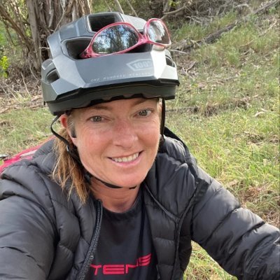A woman wearing a bike helmet and sunglasses, smiling for a selfie outdoors in a grassy area with trees in the background.