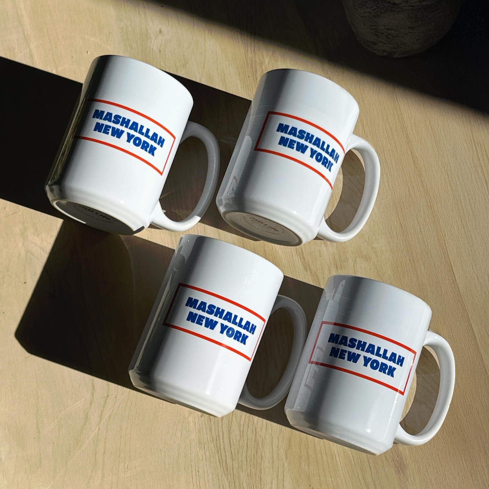 Group of white mugs with Mashallah New York text arranged on table with light shadows