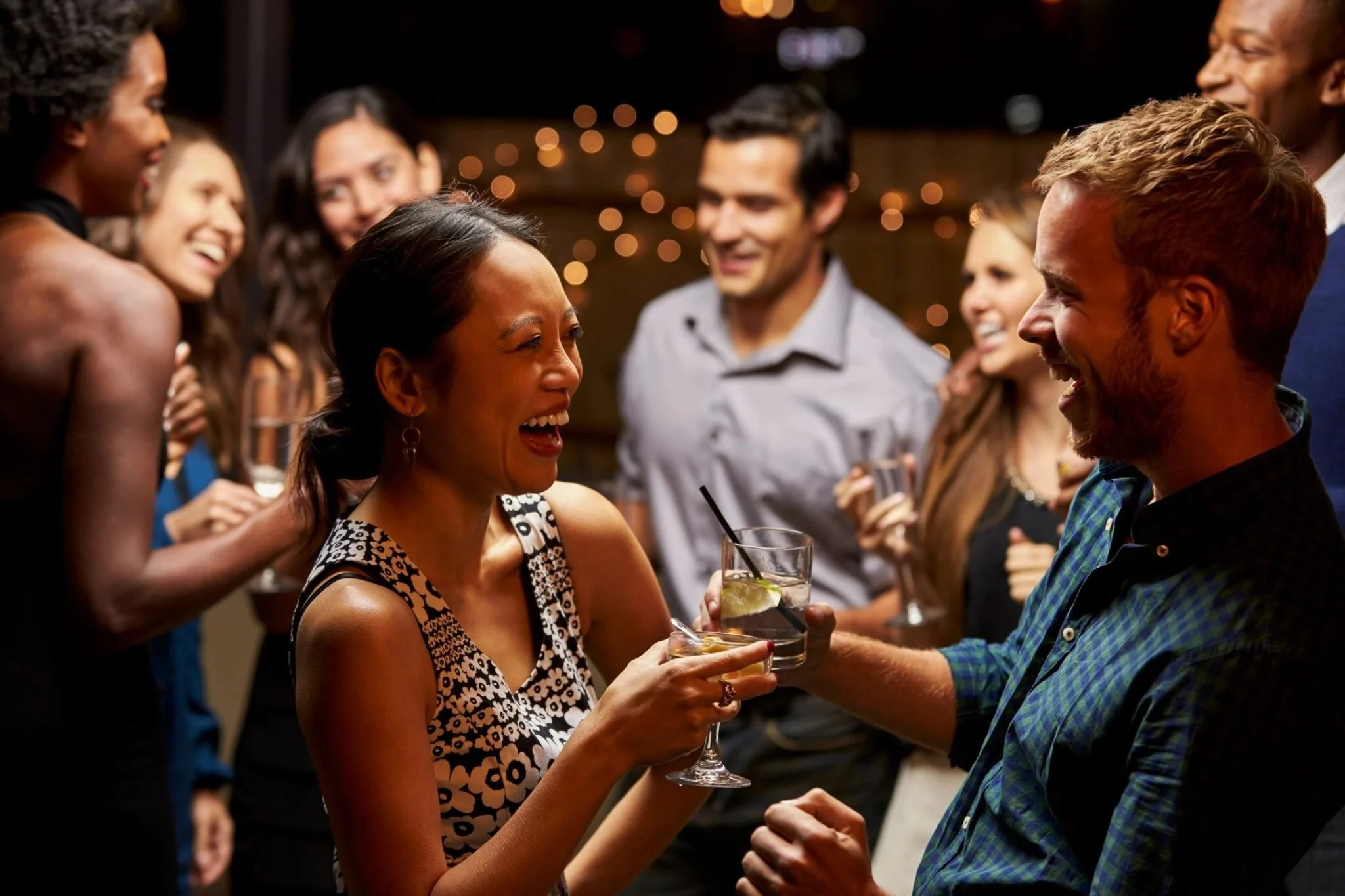 Group of people at a party, laughing and chatting with drinks in hand, in a warmly lit setting.