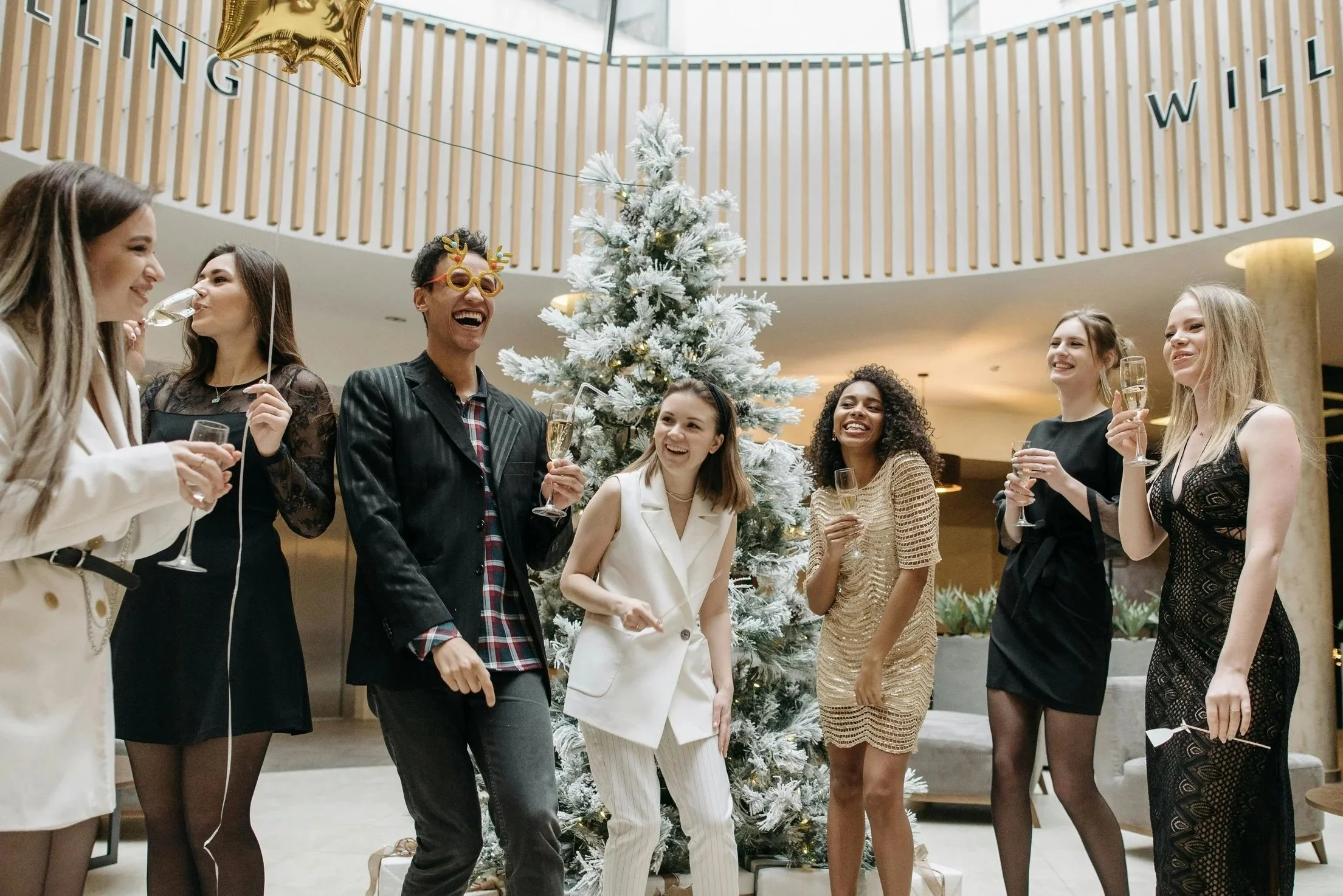 Group of seven diverse people celebrating Christmas around a decorated white Christmas tree, smiling and holding glasses of champagne in an indoor party setting.