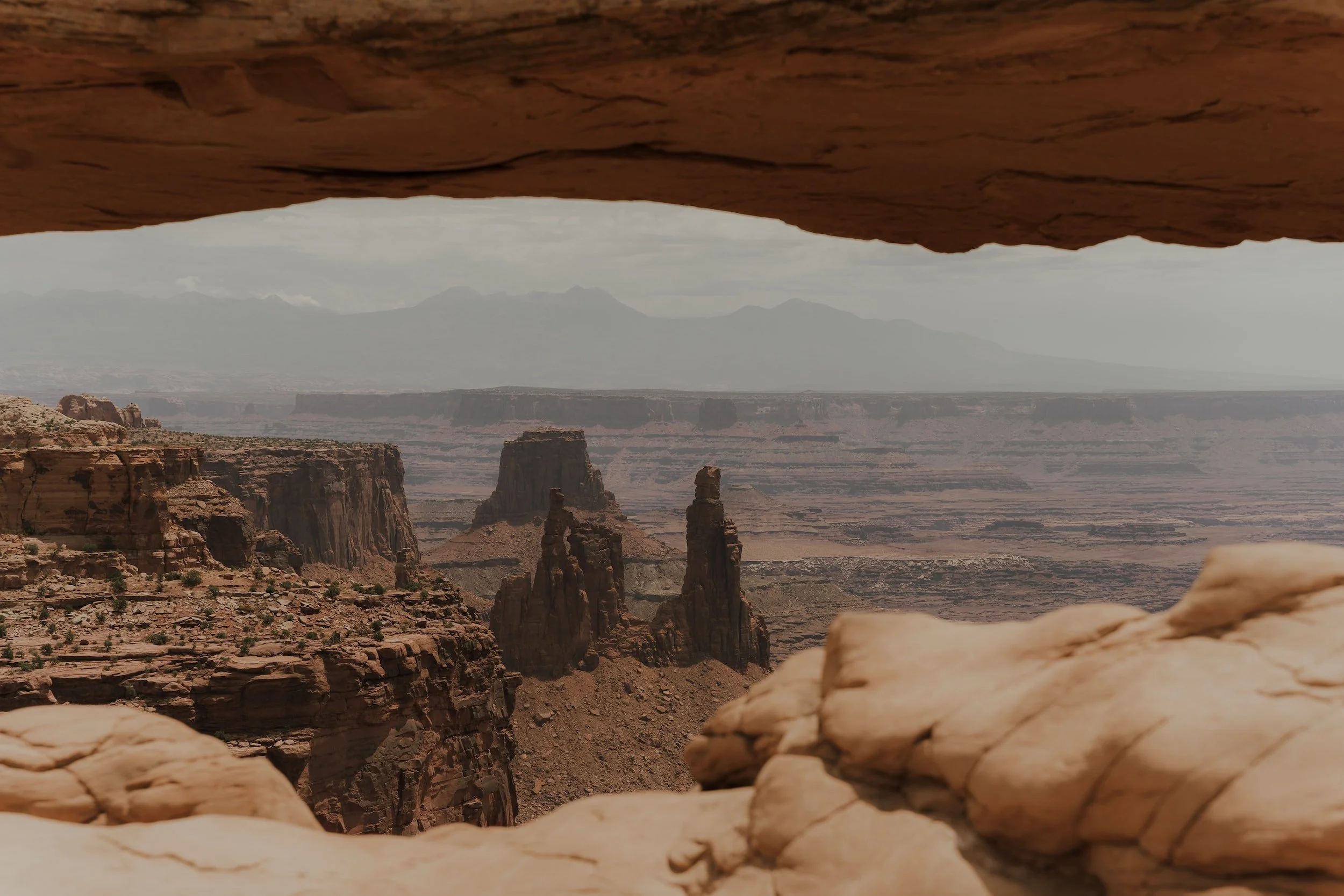 Scenic view of a canyon through a natural rock window, with layered cliffs, rock formations, and distant mountains under a cloudy sky.