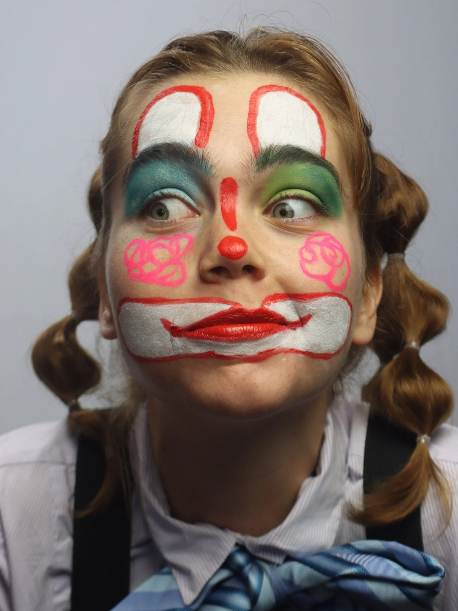 A person with clown makeup and face paint, including blue, green, pink, white, and red colors, styled to resemble a clown face.