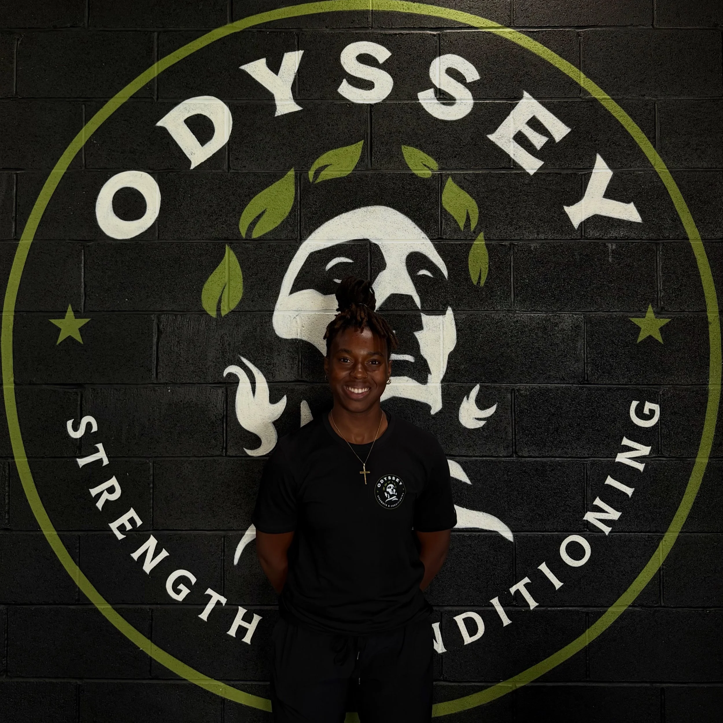 Person smiling standing in front of a black wall with a large circular logo that has skull, leaves, stars, and text.