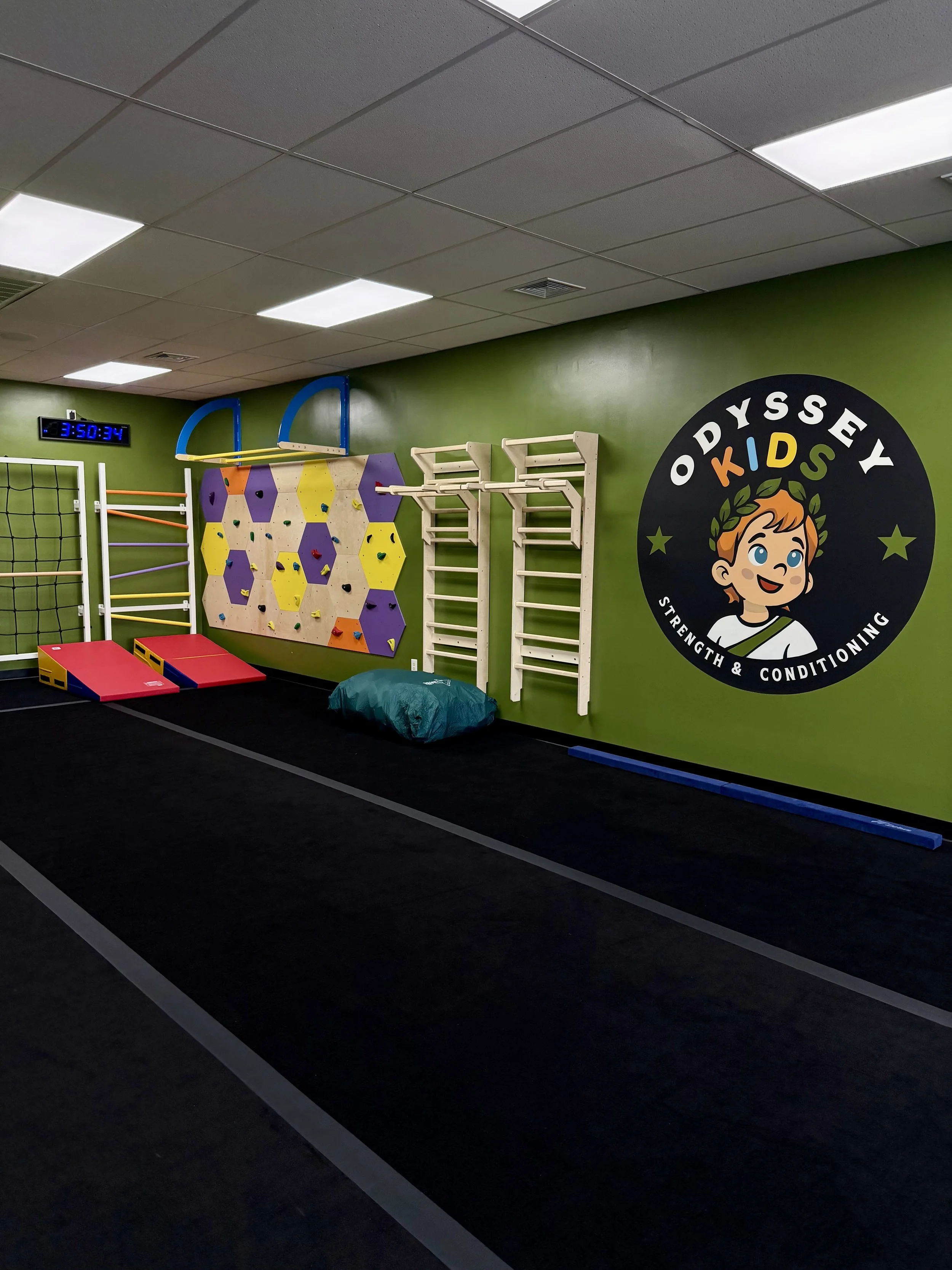 Indoor kids' play area with a climbing wall, padded mats, and gym equipment, with a large colorful sign that reads 'Odyssey Kids' on a green wall.