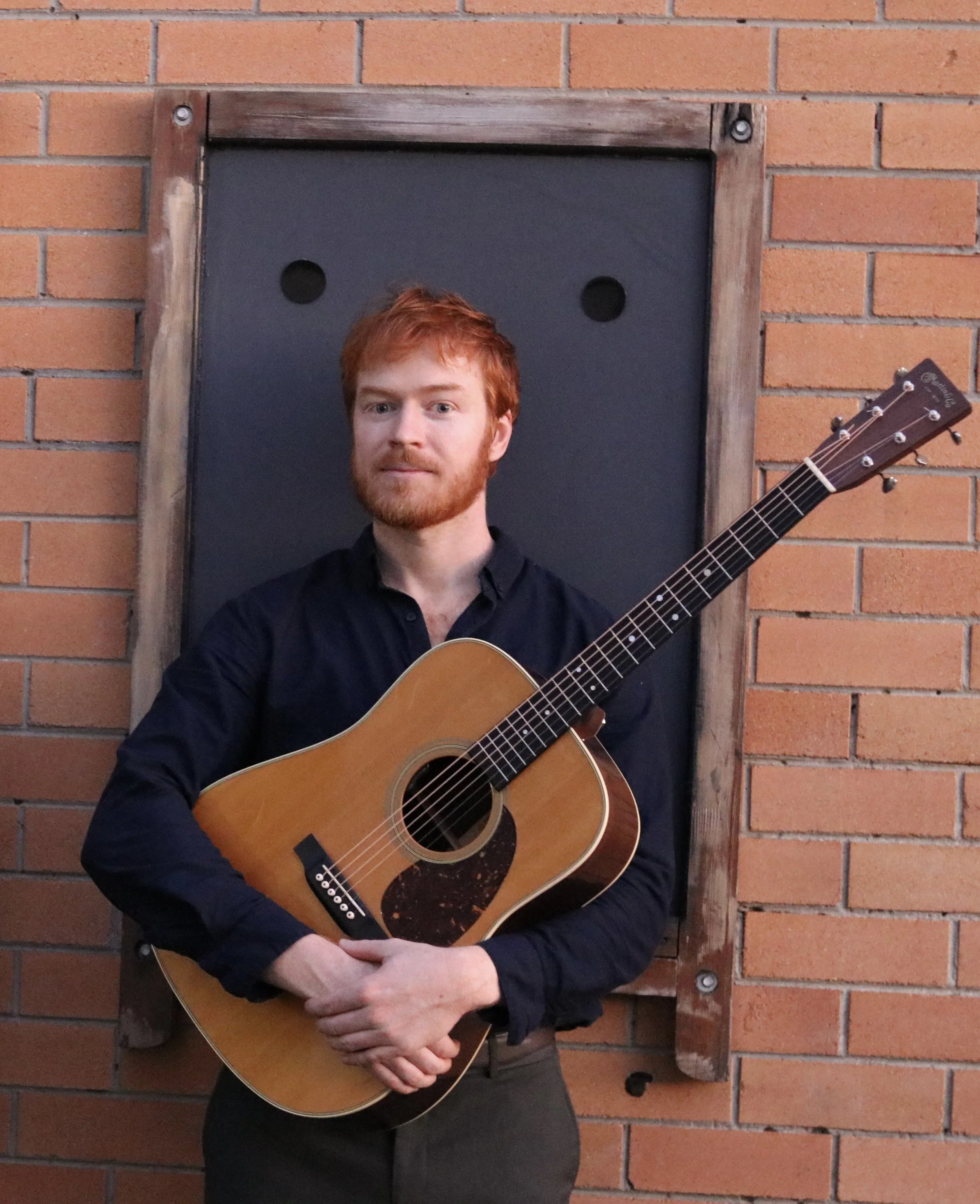 A man with red hair and a beard holding an acoustic guitar, standing in front of a brick wall with a blackboard framed with wood.