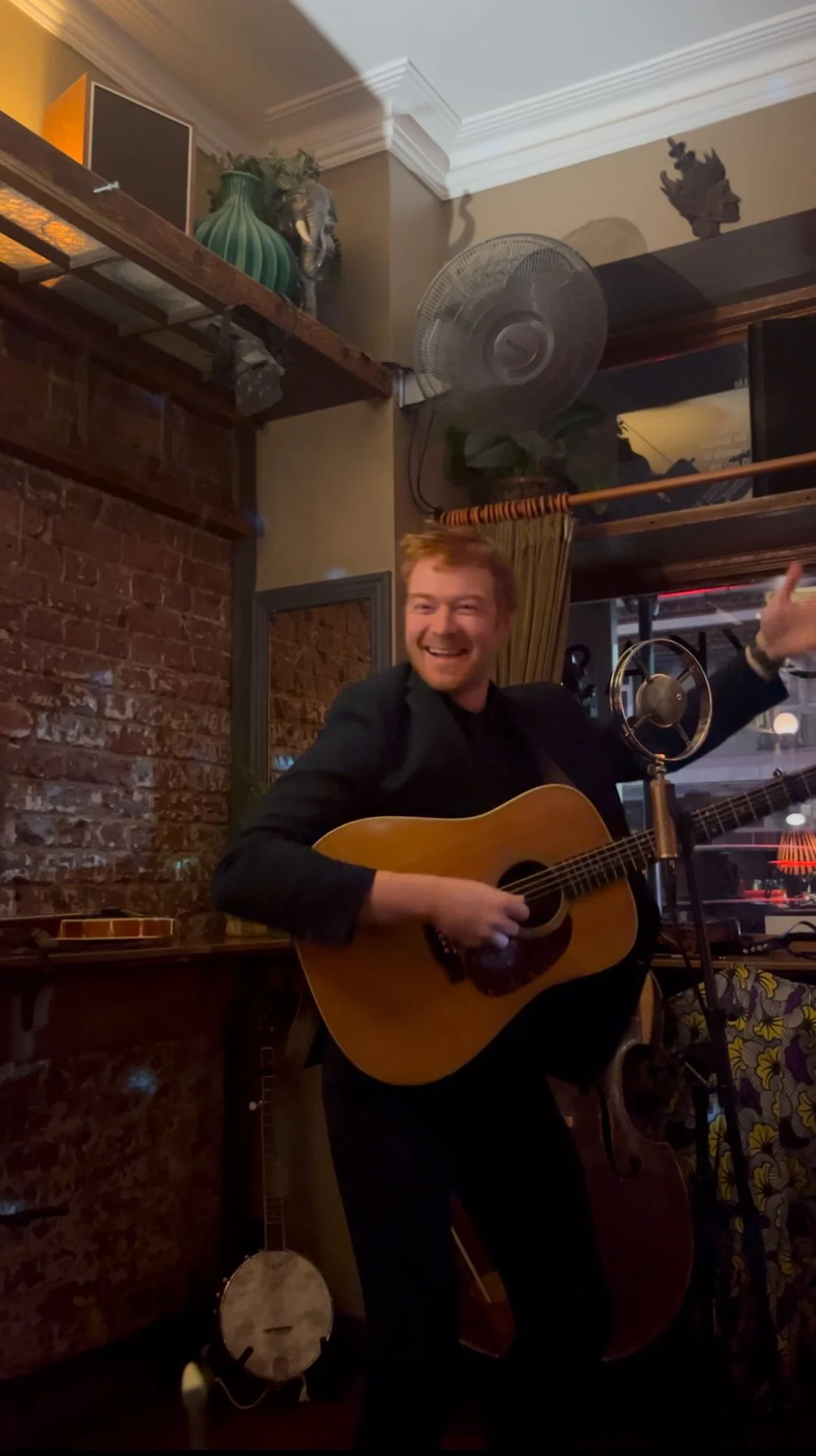 A man with red hair and a beard playing an acoustic guitar, smiling and gesturing with his right arm, in a cozy indoor setting with brick walls and musical instruments.