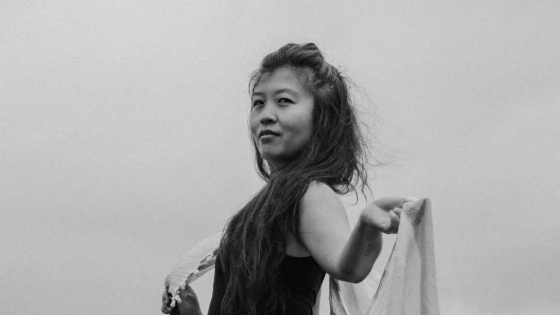 A woman with long, wavy hair holding a shawl or scarf over her shoulder, looking towards the camera in black and white.