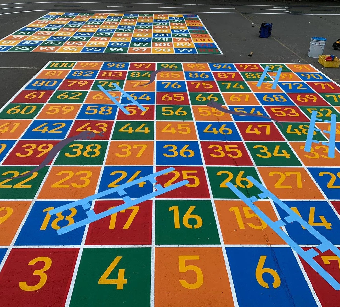 Colorful outdoor hopscotch game painted on pavement with numbered squares from 1 to 100, featuring blue plastic hurdles placed on some squares.