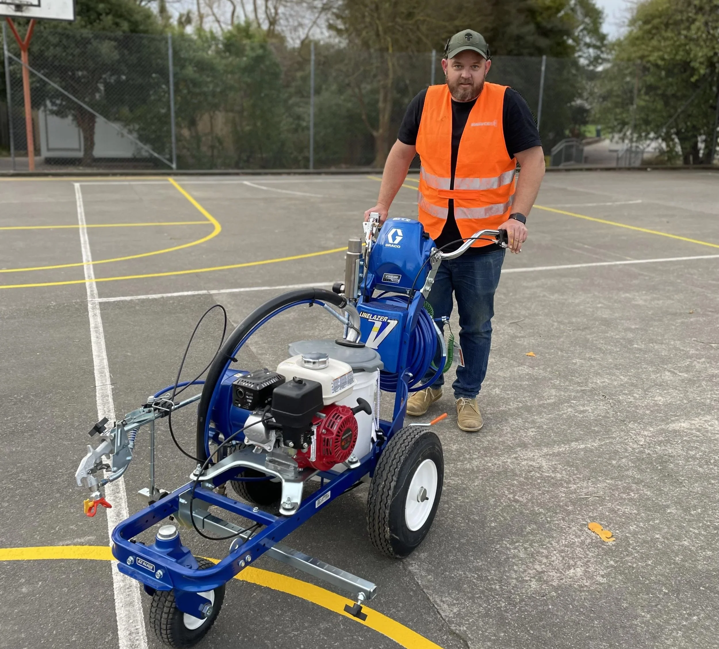 A man wearing an orange safety vest standing on a basketball court, pushing a blue LineLazer paint line striping machine.