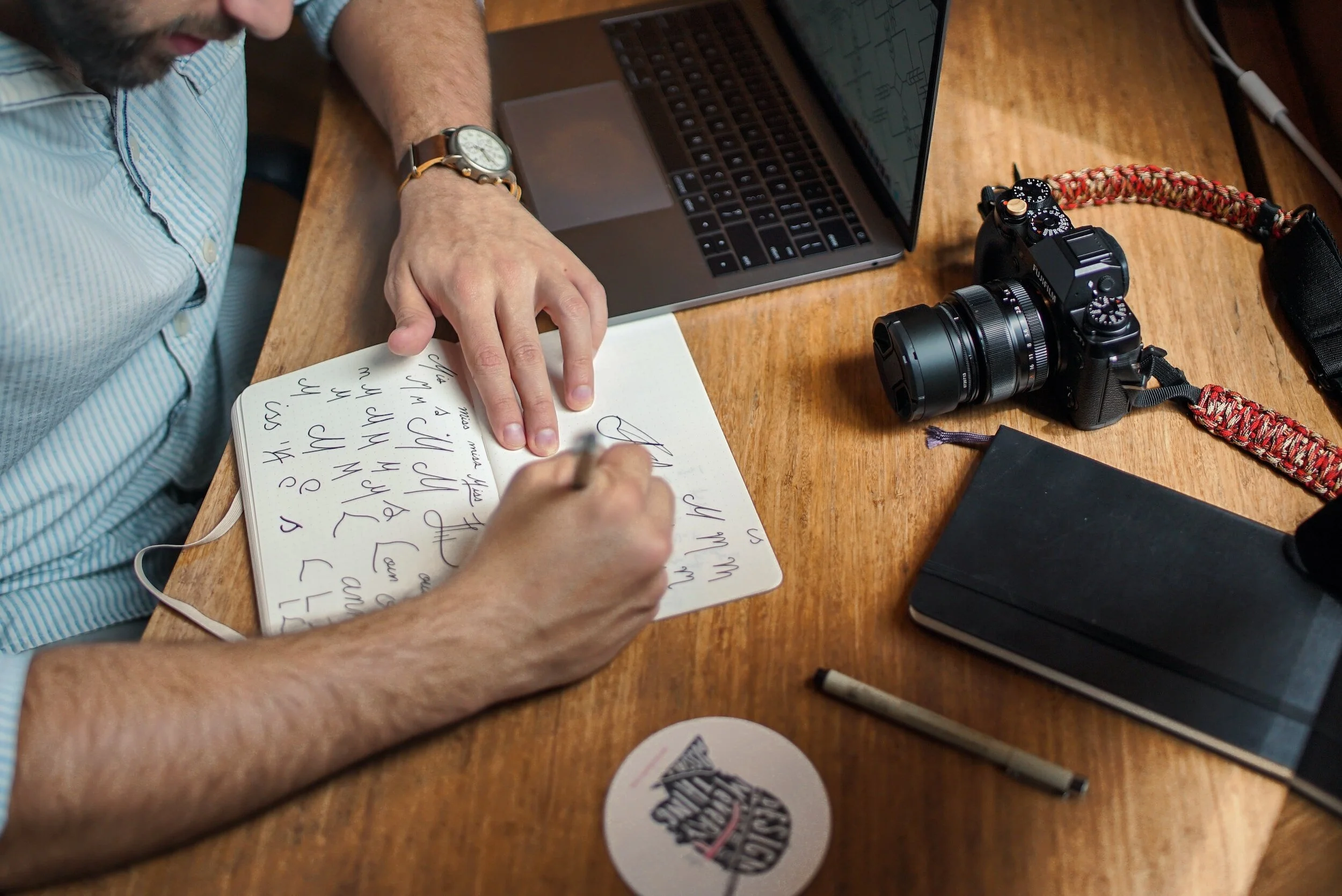 Man writing handwriting on a white notebook at a wooden table, surrounded by a camera, a laptop, and a black notebook.