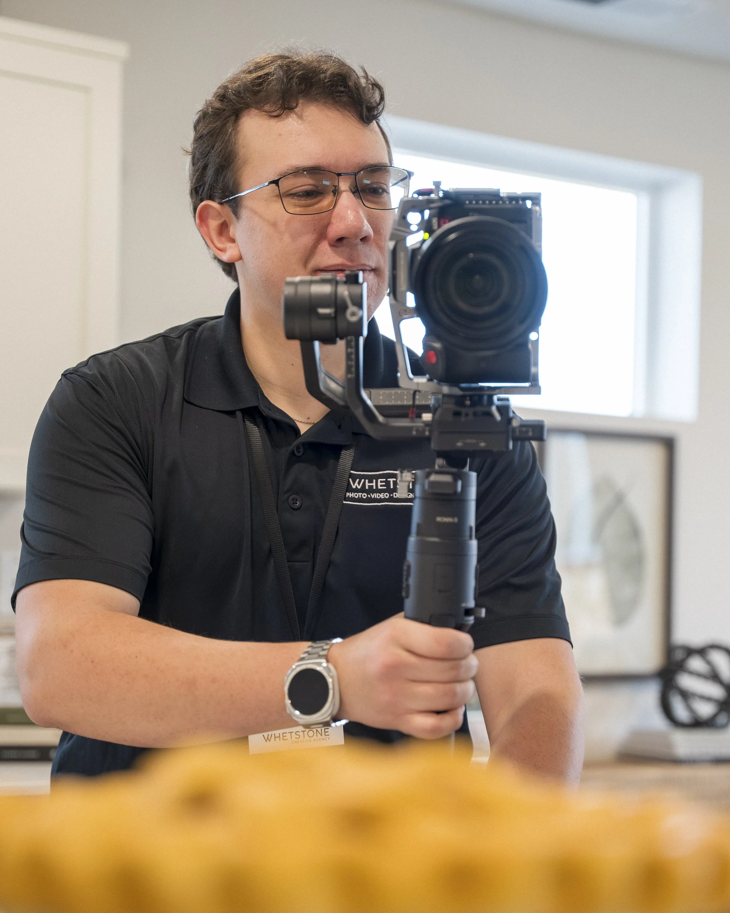 A man holding a professional camera mounted on a stabilizer system outdoors, smiling, with trees and a park bench in the background.