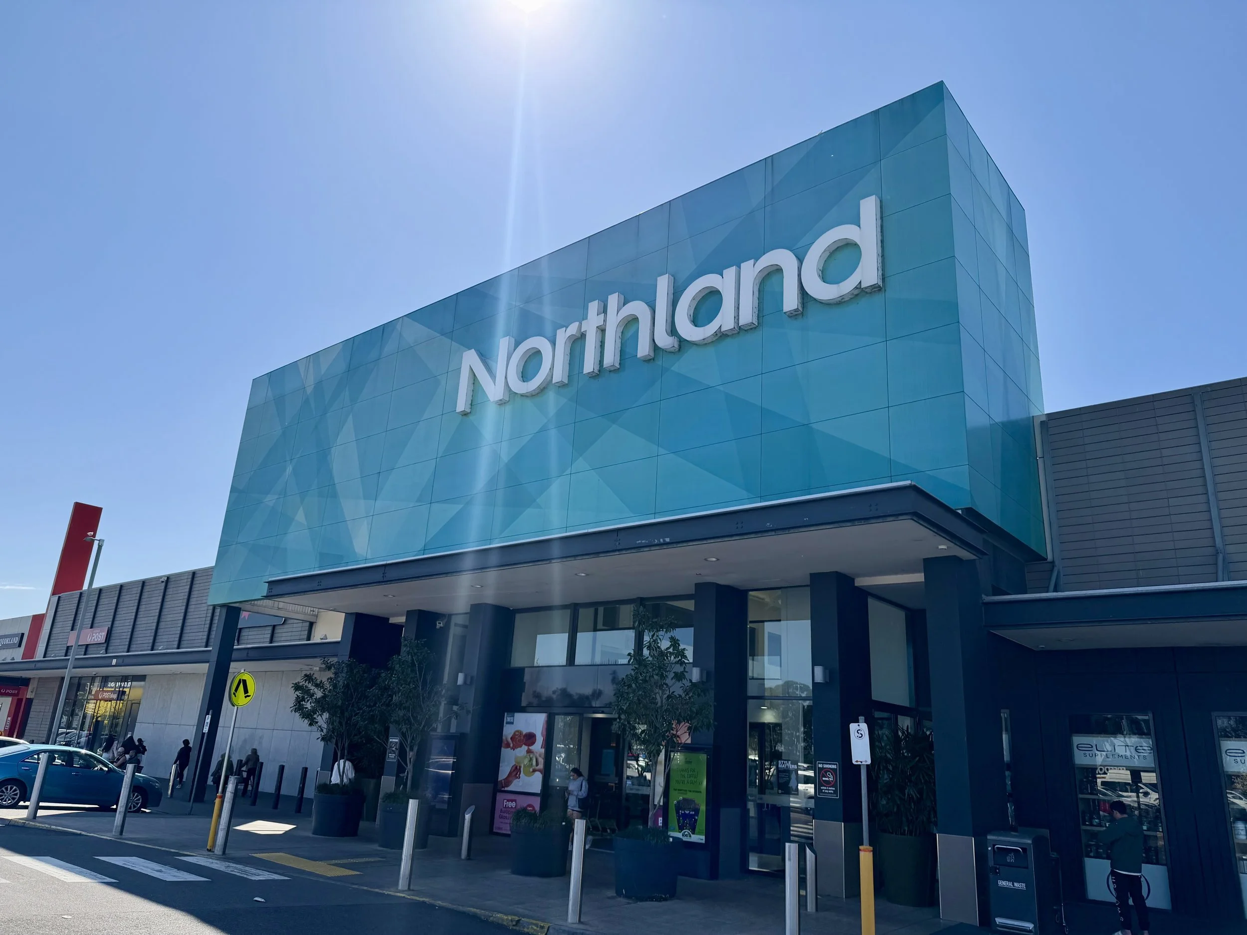 The exterior of a Northland shopping mall or retail center with a large blue sign, several people walking, and parked cars in a sunny day.