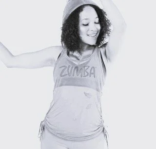 Black and white photo of a young woman smiling, wearing a sleeveless top with a graphic of lips, multiple bracelets, and a necklace, standing with hands on hips against a plain background.