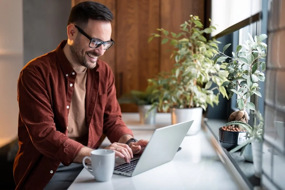 En mand arbejder på sin bærbare computer ved et vindue med planter.