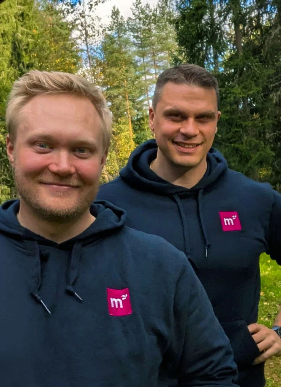 Kaksi hymyilevää miestä, jotka katsovat kameraan. Molemmilla on hupparit päällä. Miesten takana näkyy puita.