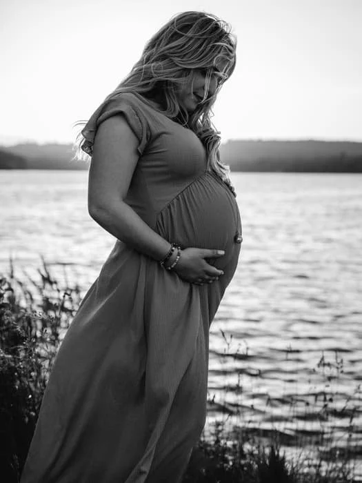 Femme enceinte en robe longue près d'un plan d'eau, avant-plan en noir et blanc.