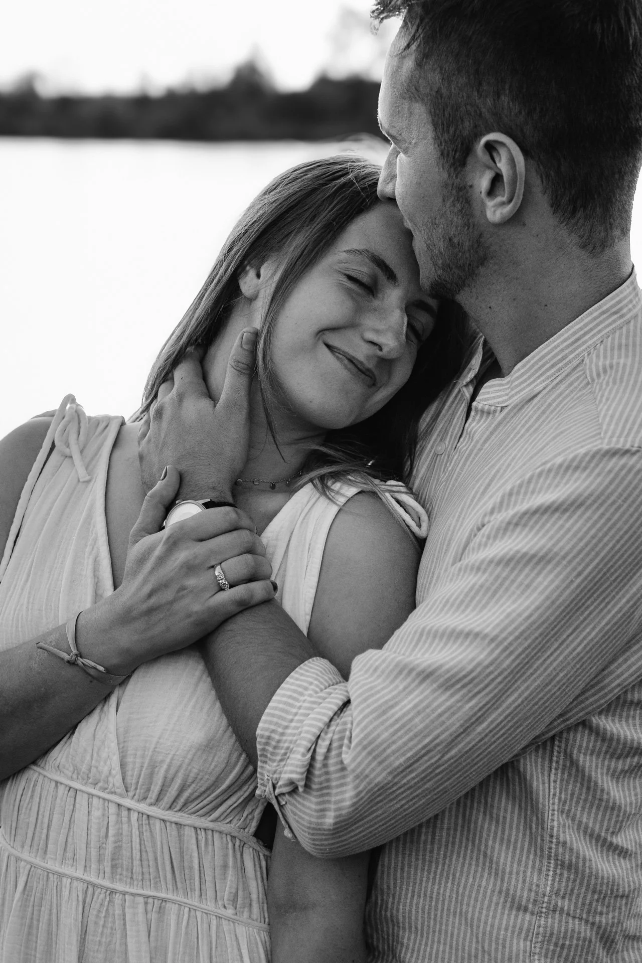 Un couple souriant et se tenant par la main près d'une rivière ou d'un lac, en noir et blanc.