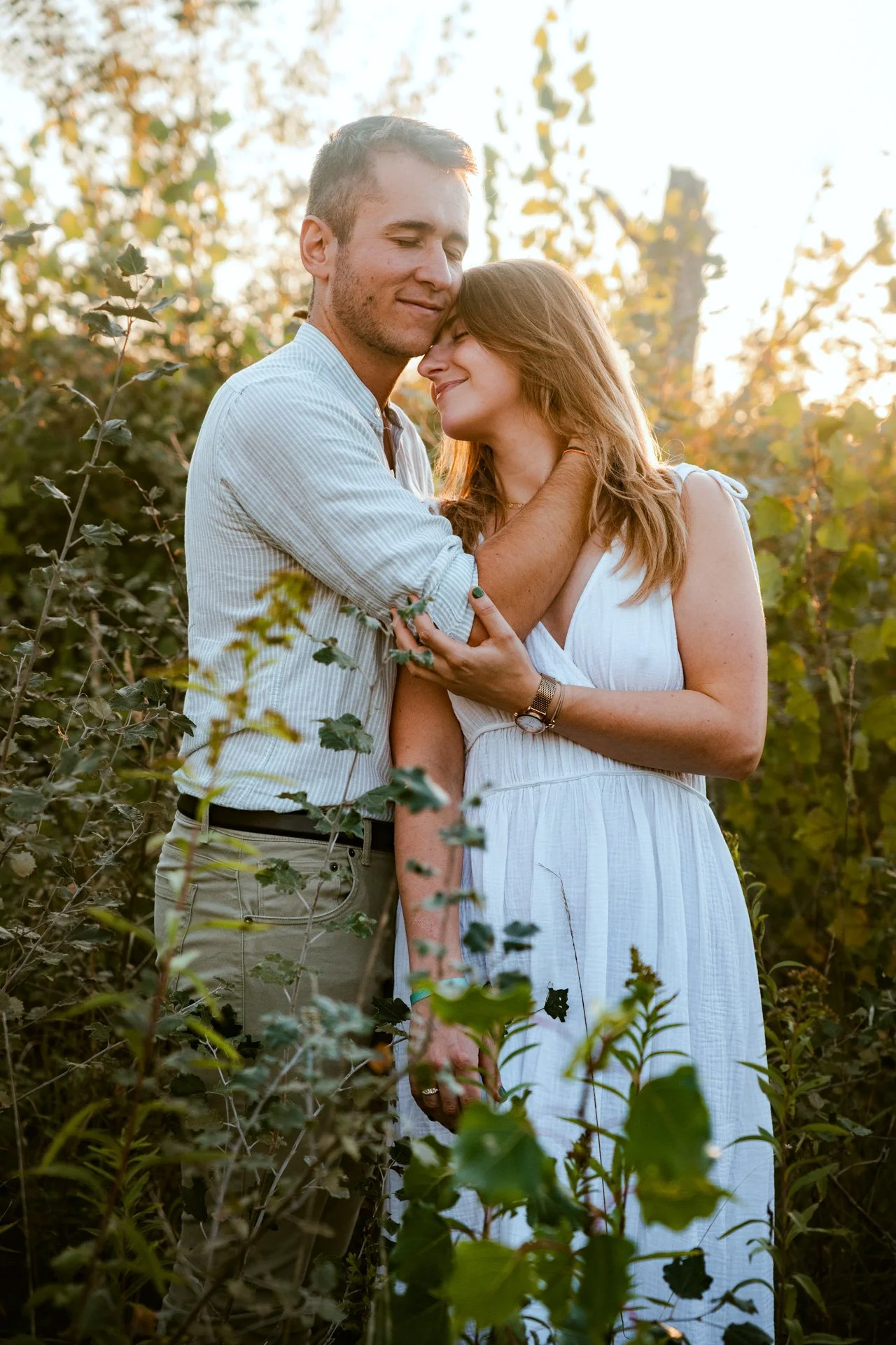 Un couple s'enlace et partage un moment intime dans un champ en plein air, avec des arbres et le soleil couchant en arrière-plan.