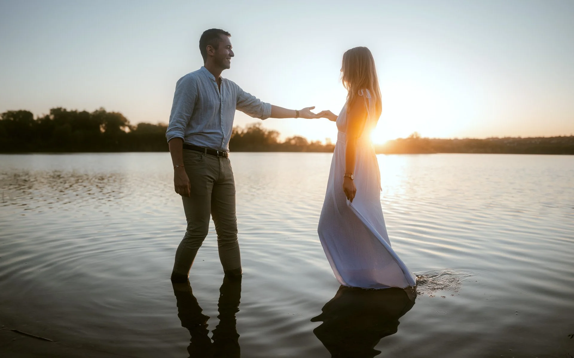 Un homme et une femme se tiennent par la main dans un lac au coucher du soleil, avec le soleil derrière eux.
