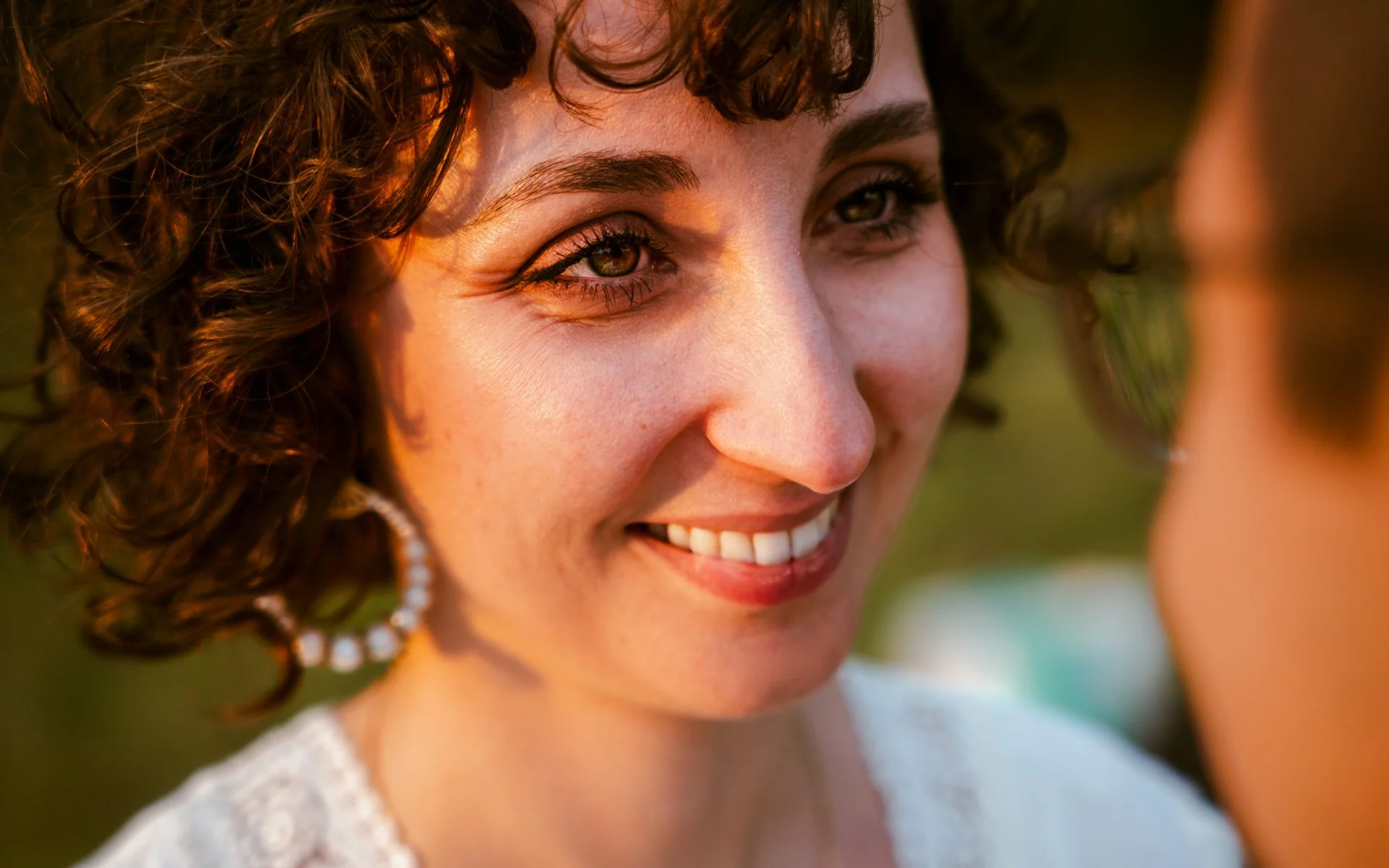 Une femme souriante avec des yeux marron, des cheveux bouclés et courts, portant une boucle d'oreille perle, en extérieur lors du coucher de soleil.