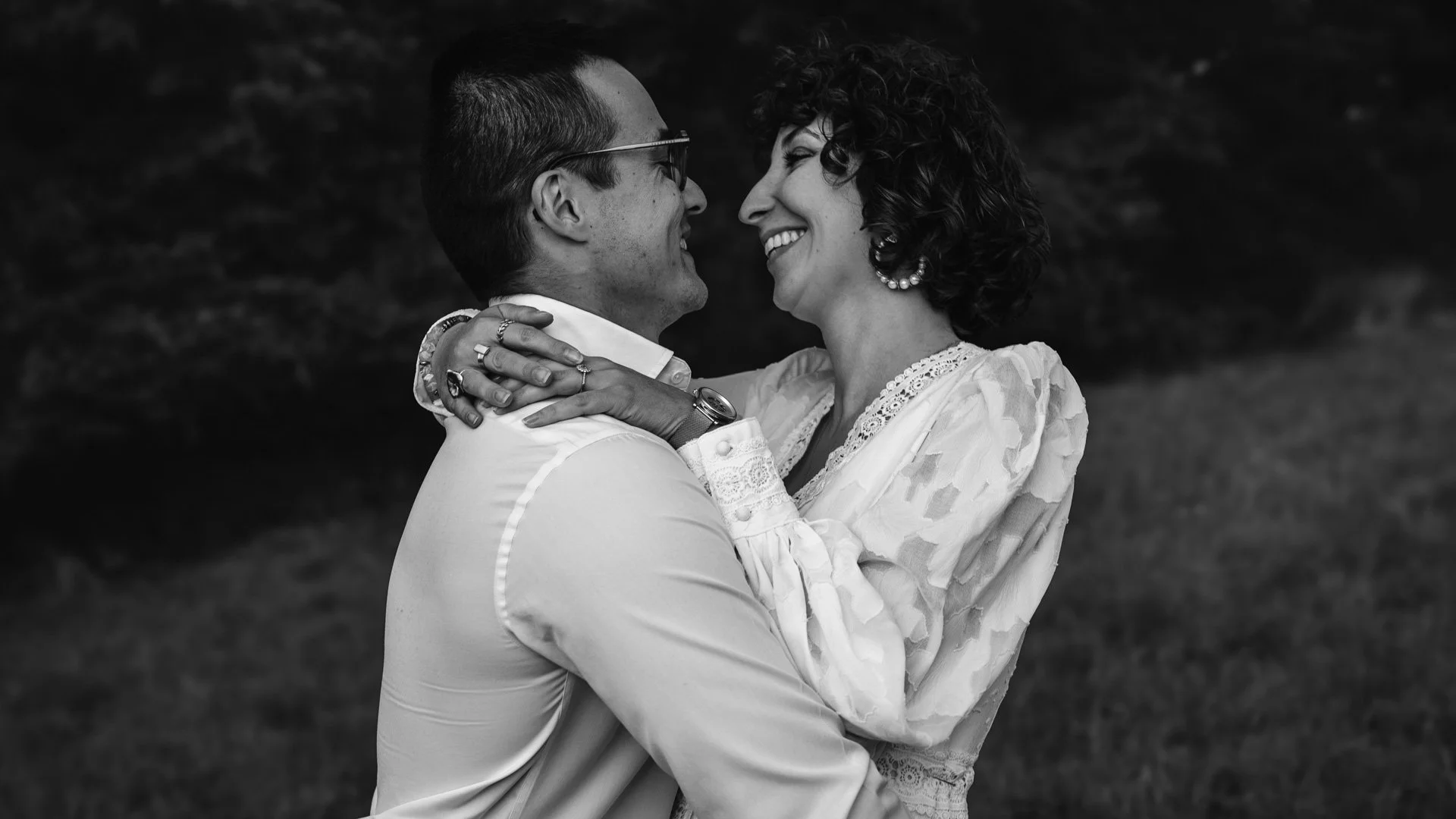 Un couple souriant et regardant dans les yeux, se tenant dans une étreinte romantique en extérieur, en noir et blanc.