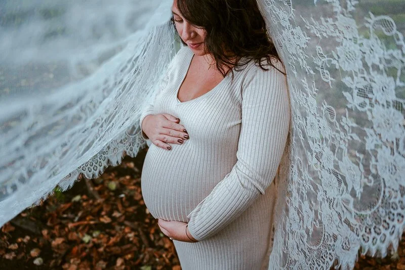 Femme enceinte portant une robe beige, tenant son ventre, debout devant un voile en dentelle.