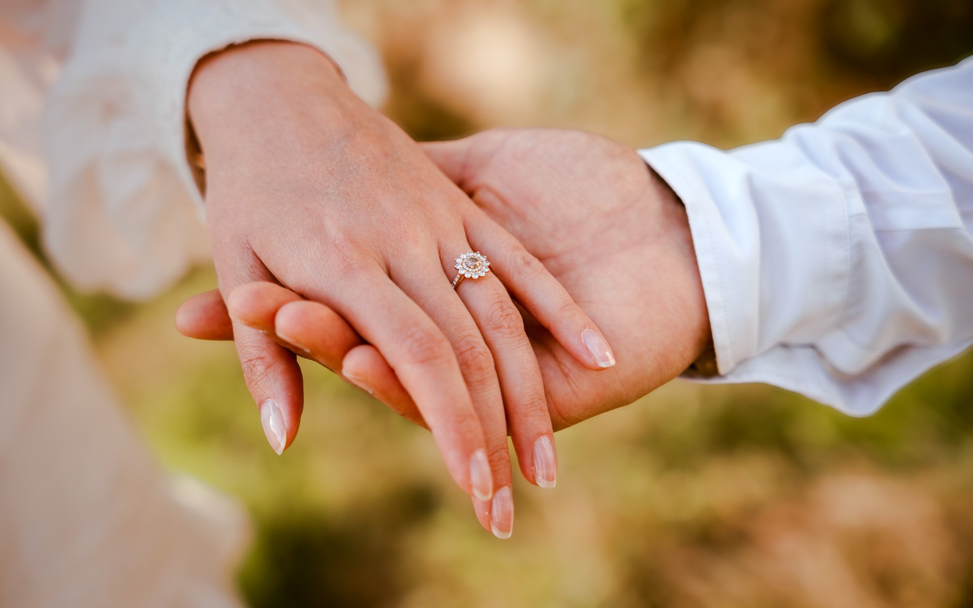 Mains de deux personnes entrelacées, une porte un anneau de mariage avec des diamants, dans un environnement naturel.