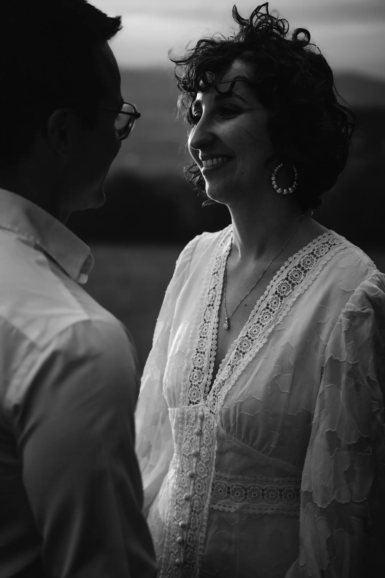 Une femme et un homme regardent l'un l'autre de près, en noir et blanc, dans un paysage rural, souriant tous les deux, lors d'un moment intime
