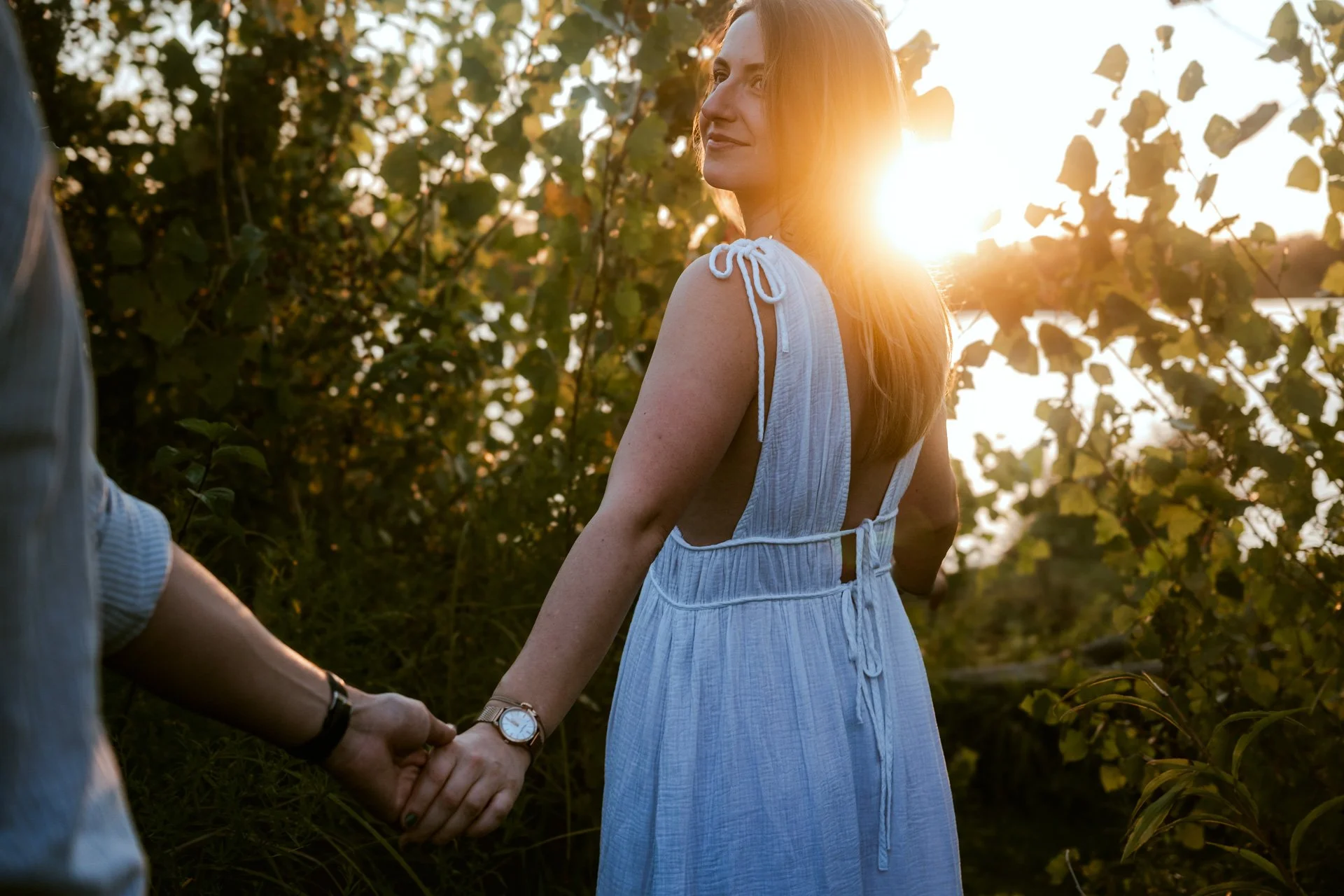 Une femme en robe blanche tenant la main d'une personne hors cadre, au coucher du soleil, dans un environnement naturel avec des feuilles vertes.
