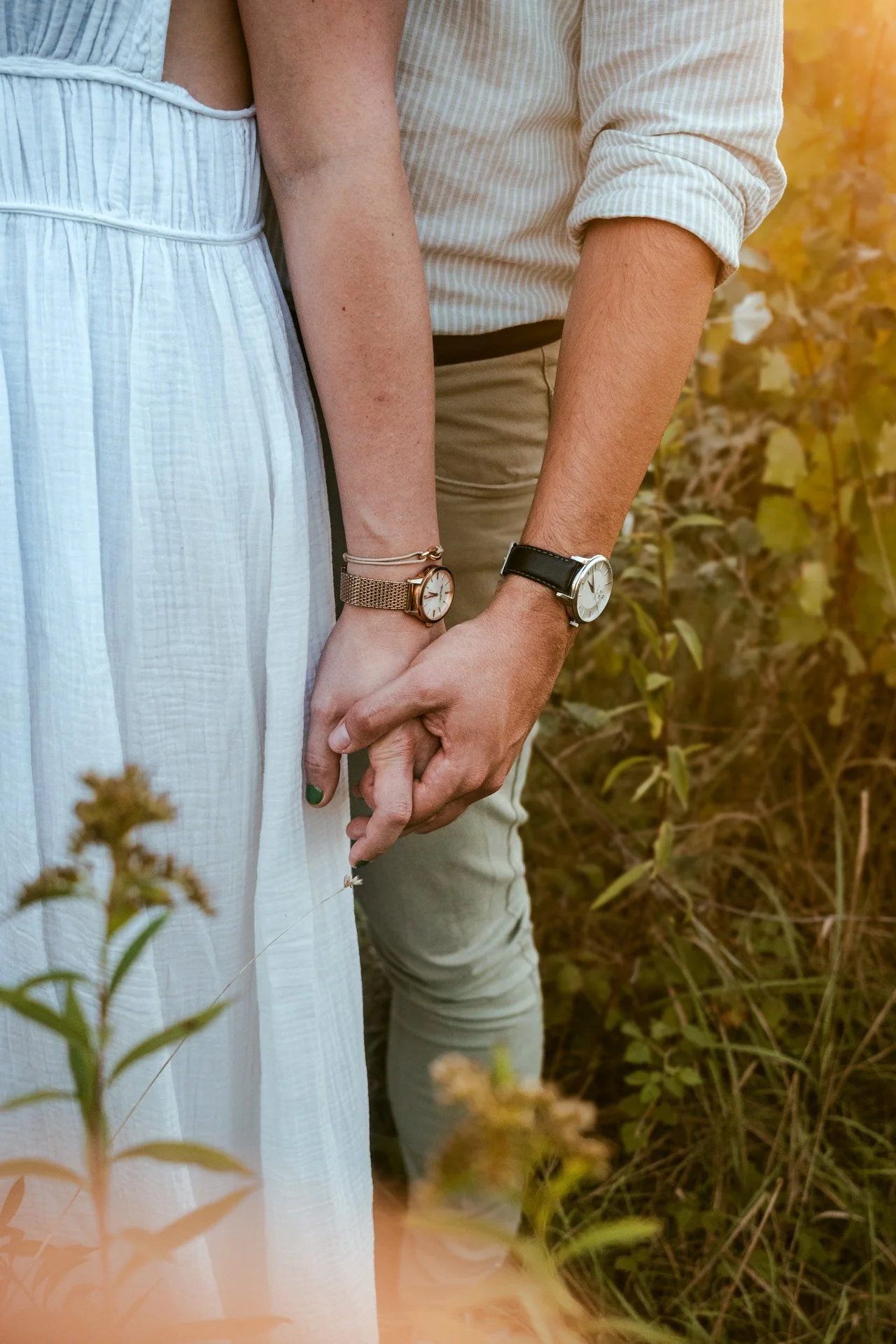 Un couple tenant leurs doigts ensemble dans un champ de végétation, portant des montres élégantes et des vêtements décontractés.